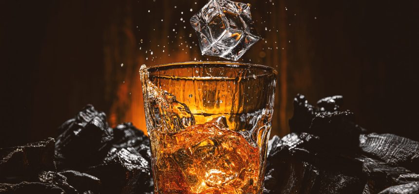 ice cubes fall into a glass with a brown alcoholic drink. splashes from falling ice fly in different directions